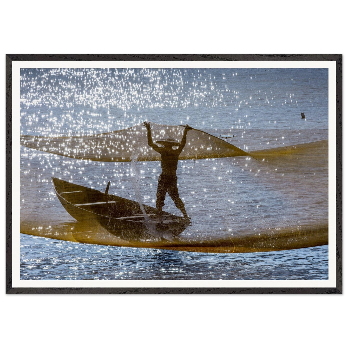 A captivating photography of a timeless moment. In a small canoe-like boat, this piece seizes the magical instant when the net is suspended mid-air, draping the fisherman in a beautiful, golden veil. Poster. Frame.
