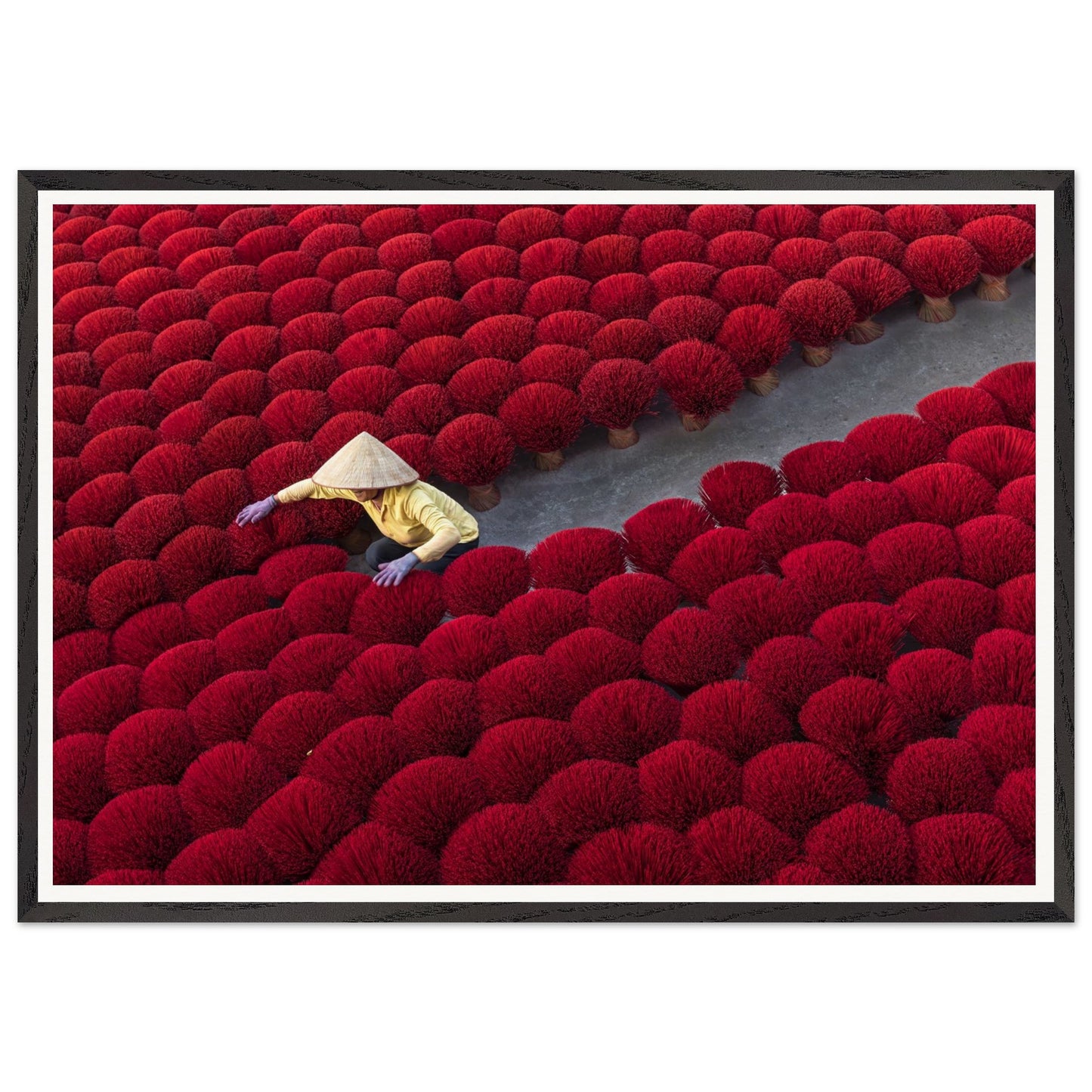 A photography of a local figure in traditional attire and a conical hat, gracefully moving through a sea of cherry red incense sticks, opening a path. Poster. Frame.
