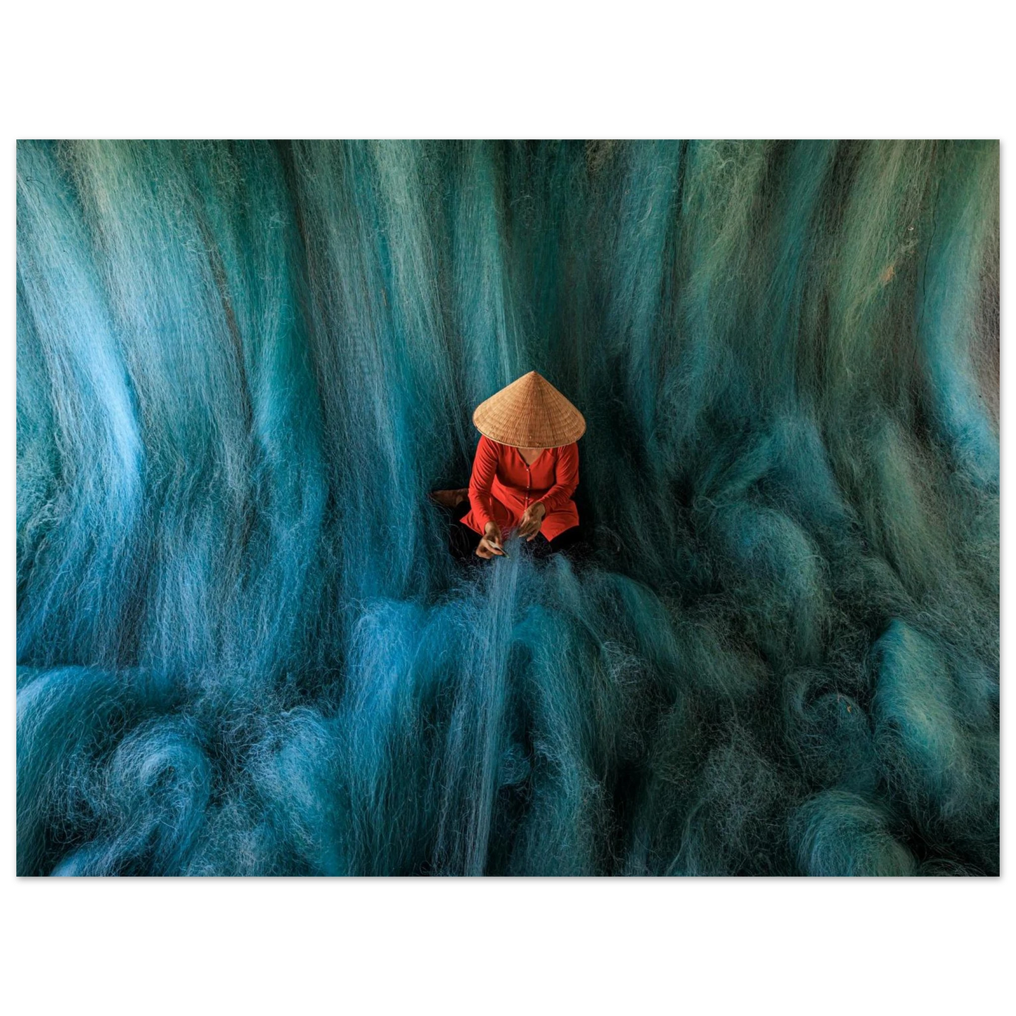 Fine art photo of a person in a conical hat mending blue fishing nets, resembling a soft waterfall flowing around them. Museum Quality printing materials.