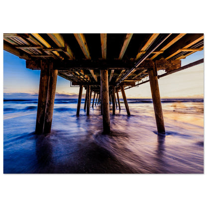 A photographic print depicting the underside of a wooden pier extending into calm waters, with a blend of light gold, blue, and purple colors representing a harmonious palette of nature and human craftsmanship.