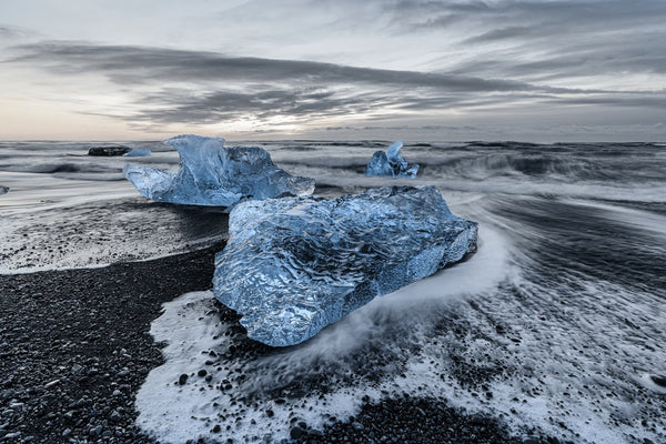Landscape photography showcases the delicate harmony between blue ice and water, seamlessly intertwined with the dramatic black stones on the beach. Poster.