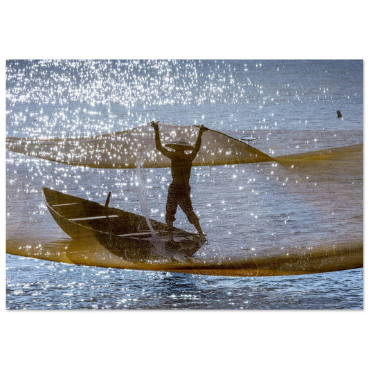 A captivating photography of a timeless moment. In a small canoe-like boat, this piece seizes the magical instant when the net is suspended mid-air, draping the fisherman in a beautiful, golden veil. Poster.