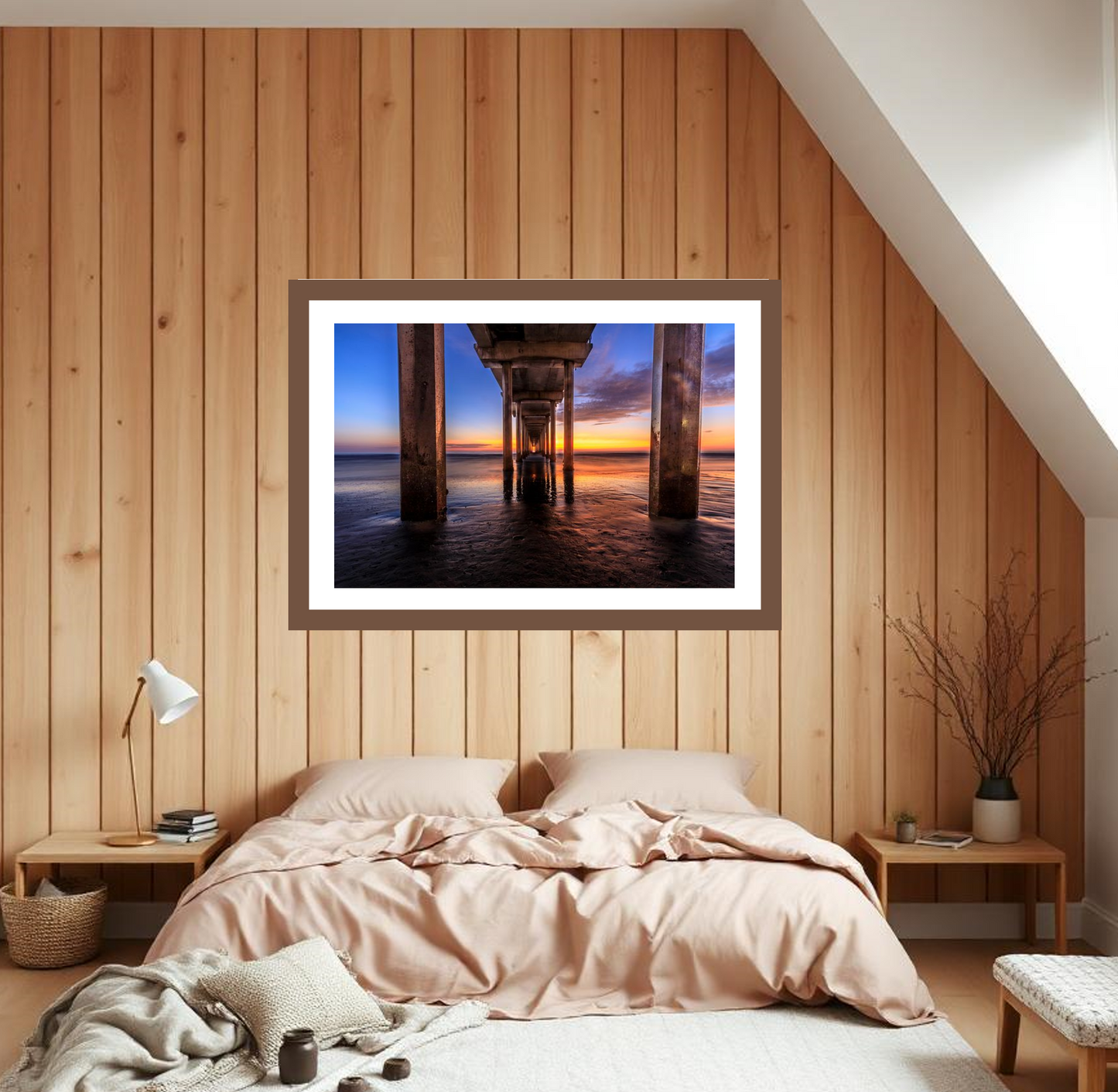 Framed artwork of a sunset under a pier above a bed with pink bedding in a bedroom with wooden walls.