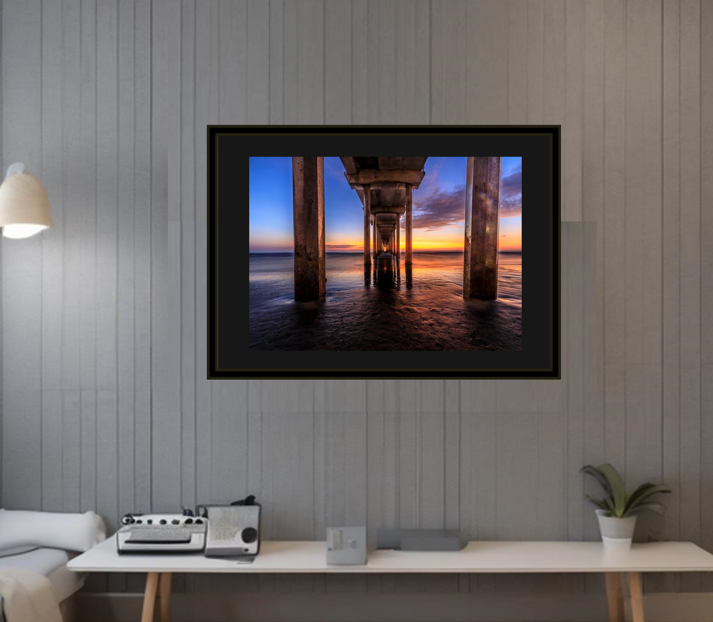 Framed artwork of a sunset under a pier on a wall in a room with a desk and lamp.