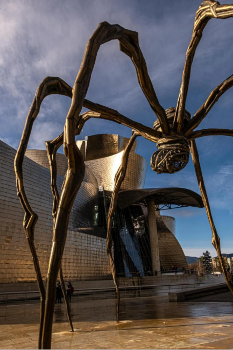A beautiful steel spider with long legs standing outside a modern steel building as the perfect sentinel.