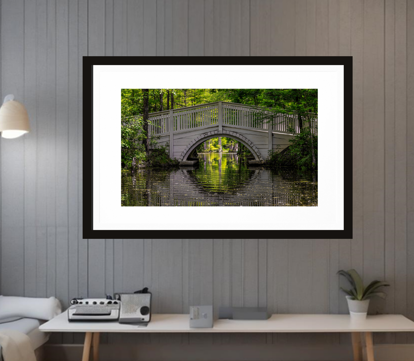 Framed photograph of a bridge over water in a forest, displayed on a wall above a desk elevating the space with decoration with soul.